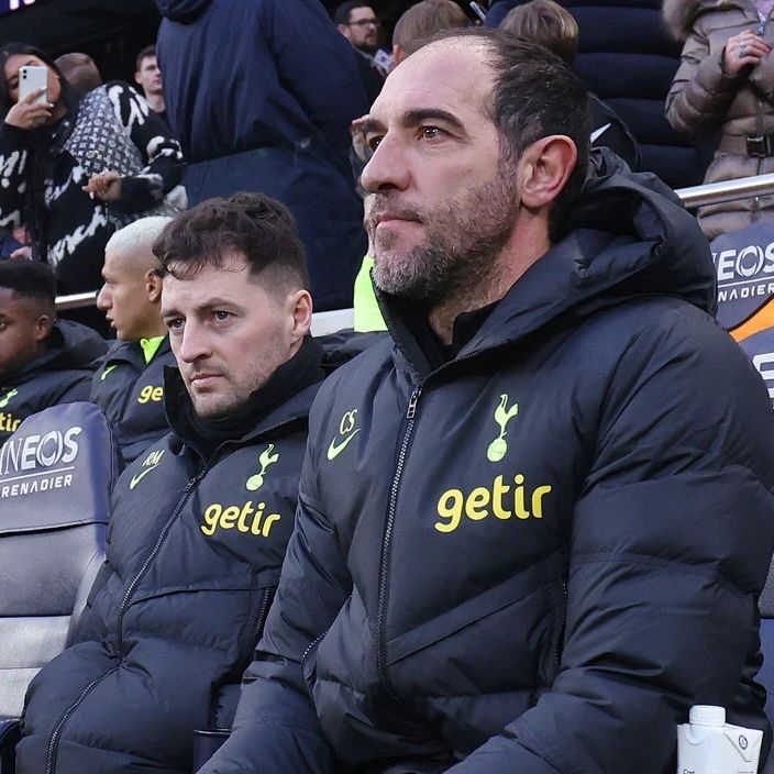 Cristian Stellini sits on the Tottenham bench in the foreground, with Ryan Mason beside him.