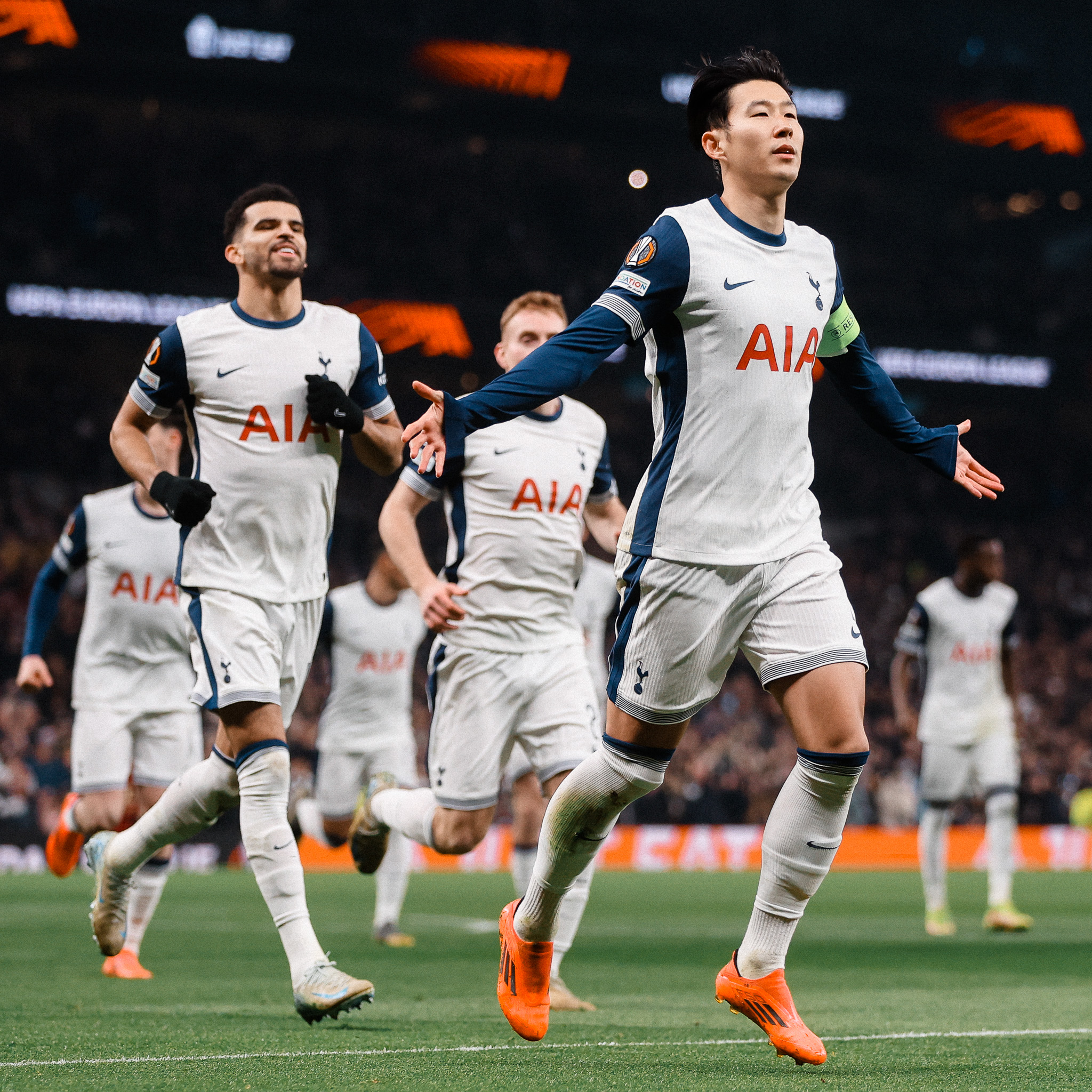 Son Heung-min stretches his arms out to celebrate scoring a penalty as his teammates run up behind him.