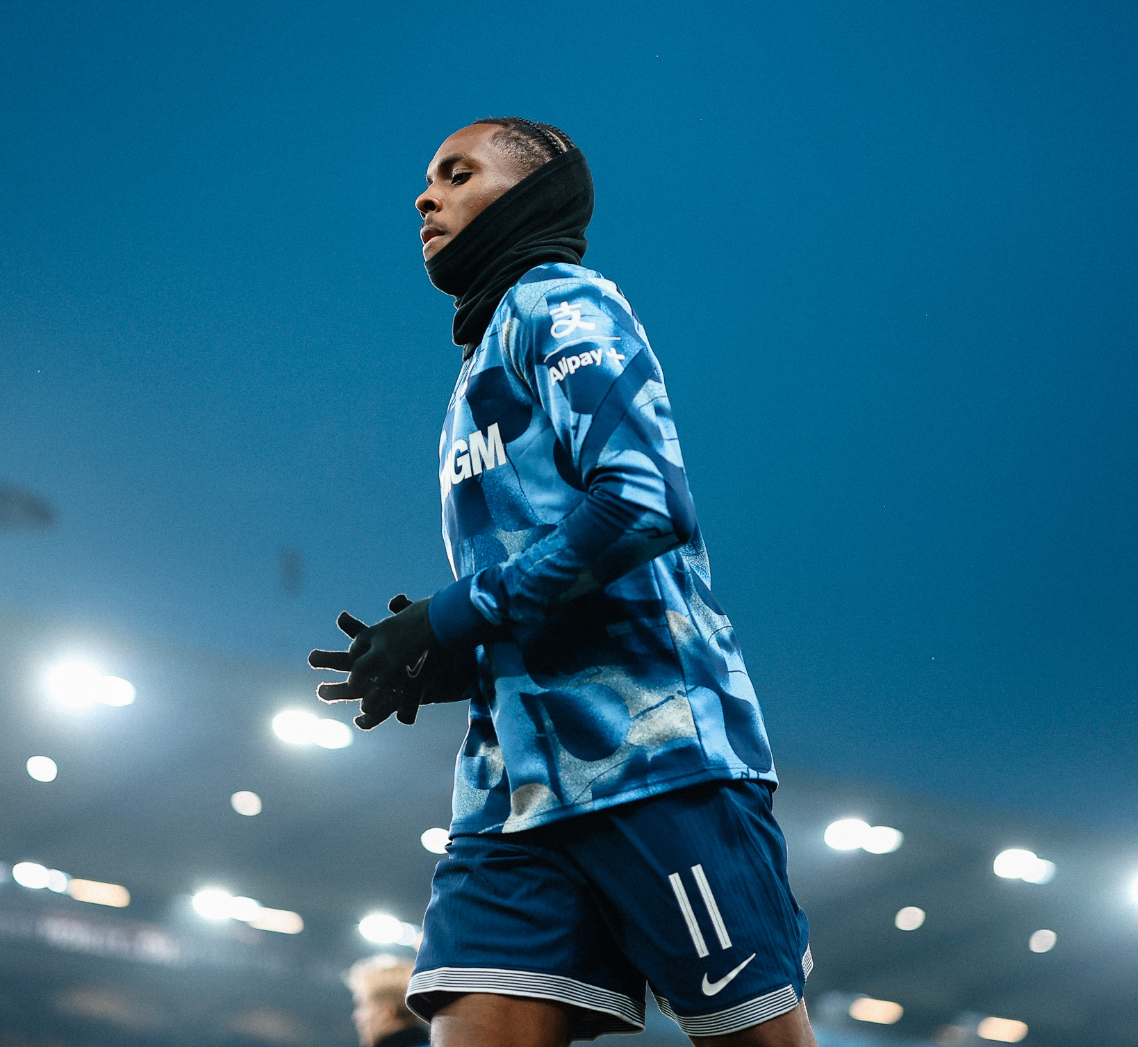 Mathys Tel warms up under the lights at Villa Park in his all-blue training wear, a snood around his neck and gloves on as well.