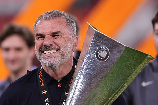 Ange Postecoglou grins toothily while holding the Europa League trophy.