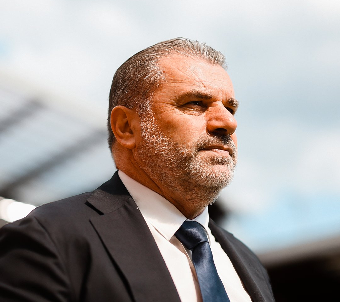a close-up shot of Ange Postecoglou in a suit, a blurred stadium in the background.