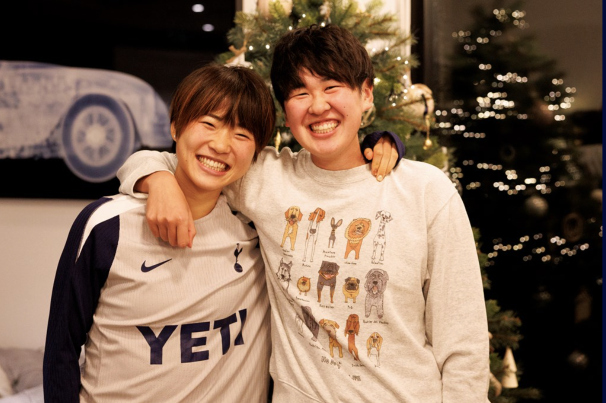 Tōko Koga puts an arm around Maika Hamano, both grinning broadly, in front of a Christmas tree.